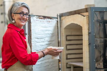 Craftswoman placing clay mugs into kiln for firing pottery while working in workshop.