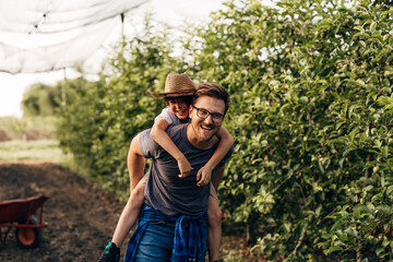 Happy Caucasian man is carrying his son on his back in the fruit plantation.