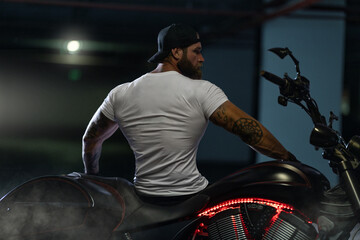 Athletic bearded biker man posing with motorbike with his back to the frame