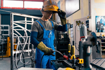 A worker is using a welding machine in his hand and working on a metal rod, he raised his visor to see better