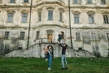 Obraz premium Mother, father throw up daughter, son into sky near old palace in Ukraine. Family spending time together in nature. Mom, dad hug happy baby kid walk in green grass. Parents embrace child on spring day
