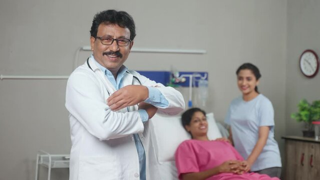Rack focus shot, confident indian doctor standing with crossed arms in front of patient by looking camera at hospital - concept of professional occupation, expertise and successful treatment