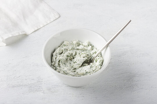 White bowl with cottage cheese cream mixed with herbs and garlic on a light gray background. Cooking appetizer or toppings for stuffing vegetables, vegetarian food