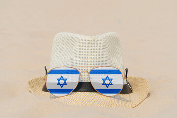Fototapeta na wymiar Sunglasses with glasses in the form of the flag of Israel and a hat lie on the sand. The concept of summer holidays, travel and tourism in Israel