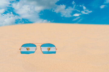 Sunglasses with glasses in the form of the flag of Argentina lie on the sand against the blue sky. The concept of summer holidays, travel and tourism in Argentina