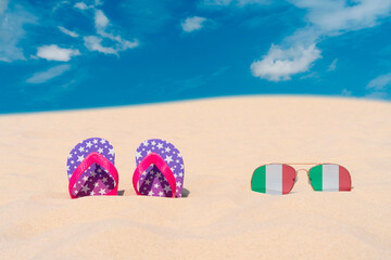 Sunglasses with glasses in the form of the flag of Italy and flip-flops lie on the sand against the...