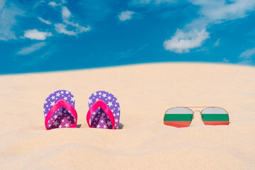 Sunglasses with glasses in the form of the flag of Bulgaria and flip-flops lie on the sand against the blue sky. The concept of summer holidays, travel and tourism in Bulgaria