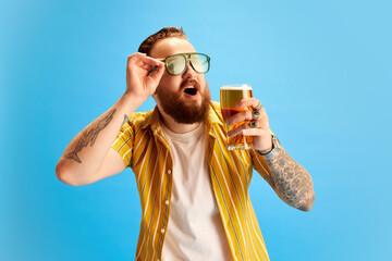 Bearded cheerful man in yellow shirt and sunglasses drinking beer against blue studio background. Vacation and relaxation. Concept of emotions, leisure time, positivity, party and celebration, ad