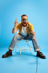 Bearded man in casual clothes and sunglasses emotionally playing console against blue studio background. Evening fun. Concept of emotions, leisure time, positivity, online video games, ad