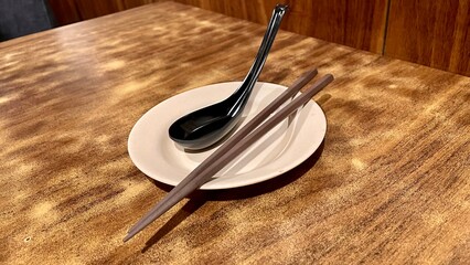 black plastic spoon and a brown wood chopstick put on a white ceramic plate on wooden table, overhead view.