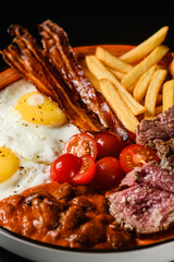Fried eggs with bacon, tomatoes, stewed beans, french fries on black background.