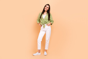 Fototapeta na wymiar Full size photo of gorgeous good mood girl dressed green blouse white trousers hold arms in pockets isolated on beige color background