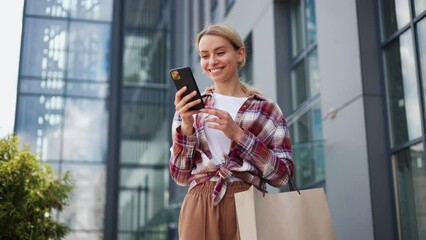 Happy excited young woman with packages receiving discount for shopping while checking email box on mobile tapping message receive cashbask sharing sales news walking in urban street shopping mall.