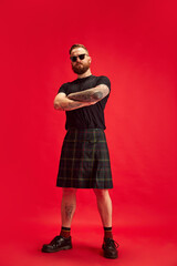 Portrait of bearded serious man in sunglasses posing in scottish skirt - kilt against red studio background. Extraordinary look. Concept of lifestyle, scottish style, fashion, music, fun and joy, ad