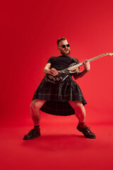 Rock and roll concert. Bearded man in sunglasses and skirt, kilt playing guitar against red studio background. Concept of lifestyle, scottish style, fashion, music, fun and joy, ad