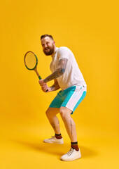 Full-length image of bearded man in sportswear posing with tennis racket against yellow studio background. Ready to play. Concept of sport, strength, fashion, emotions, lifestyle, ad