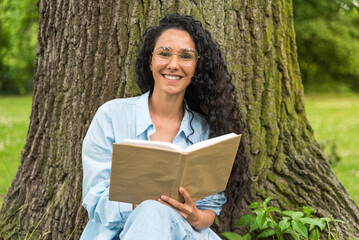 Young beautiful Middle Eastern woman with long curly hair with glasses is reading book against backdrop of tree and green city park. Portrait of a bright smiling handsome girl sitting on the grass