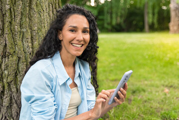 Young beautiful woman of Middle Eastern appearance in casual clothes sits in a city park holding a phone in her hands and texting. Horizontal portrait of a curly smiling girl who looks at the camera.