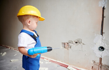 Baby boy construction worker with toy electric drill looking at the wall in apartment. Kid in safety helmet and work overalls holding cordless power tool for repair works. Home renovation concept.
