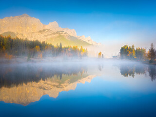 Fototapeta na wymiar Fall. A foggy morning during dawn. Autumn trees on the river bank. Mountains and forest. Reflections on the surface of the lake. Banff National Park, Alberta, Canada.