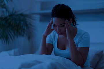 Sleepless black woman sitting in bed alone at night, touching her head
