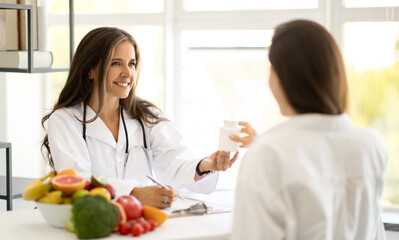 Cheerful mature caucasian lady doctor nutritionist in white coat advises young woman jar of vitamin...