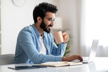 Distance Work. Handsome Indian Male Freelancer Drinking Coffee And Working With Laptop
