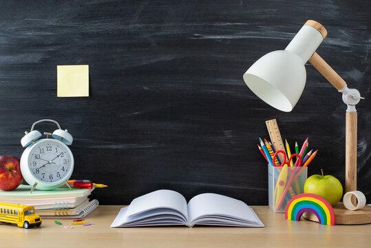 Academic setup. Side view photo of desk with stationery stand, diaries, open book, laptop, lamp, bus toy, alarm clock and school-themed props on a chalkboard backdrop. Great for text or ad placement