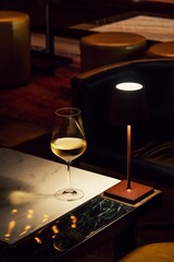 Table lamp lit brightly on a wooden table beside a stemmed glass of white wine