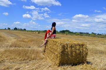 Naklejka na ściany i meble A girl with a jug in her hands sat down by a bale of hay in a mowed field on a sunny day.