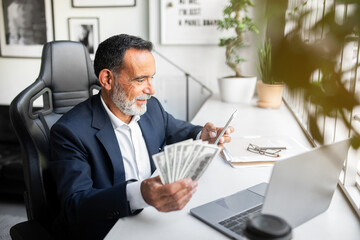 Positive caucasian senior businessman in suit use phone, computer, hold lots of money dollars in office interior