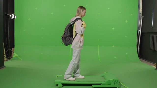 Horizontal shot on green screen background. VR or AR online walk or sport. A beautiful girl with a backpack walks in augmented reality and clicks invisible buttons. Walking on a treadmill side view.