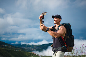 Tourist athletic man taking photo with smartphone and selfie stick on beautiful sky and fog background. Asian traveler blogger male with backpack enjoying shooting video on the top of mountains. © Pun