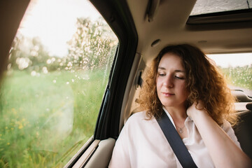 Cheerful positive curly young woman in casual wear sitting in automobile backseat with fastened seatbelt and looking away.