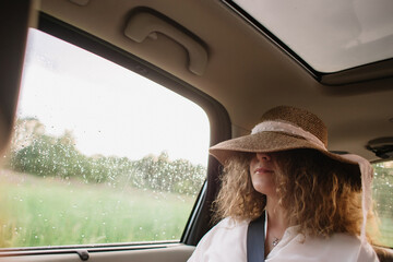 Cheerful positive curly young woman in casual wear sitting in automobile backseat with fastened seatbelt with hat.