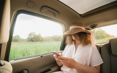 Cheerful positive curly young woman in casual wear with hat sitting in automobile backseat with fastened seatbelt and using phone.