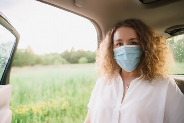 Stylish young carly woman in medical mask sitting on back seat of car on blurred background with sunset