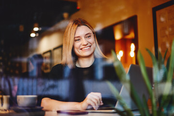 Cheerful young good looking girl sitting at a cafe and drinking coffee, smiling and using laptop 