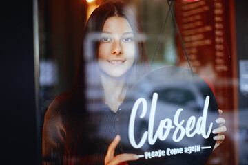 Young manager girl changing a sign from open to closed sign on door cafe looking outside waiting for clients after lockdown
