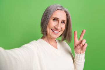 Closeup selfie blogging retired old lady wear white pullover show v-sign have fun recording her vlog isolated on green color background