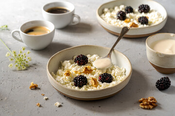 Cottage cheese in ceramic plates with blackberries, cream or yogurt, nuts and cups of coffee aside over grey concrete background