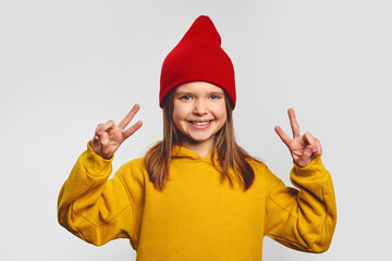 Cute girl shows victory sign with both hands, looks positively, wears yellow hoodie and red hat,...