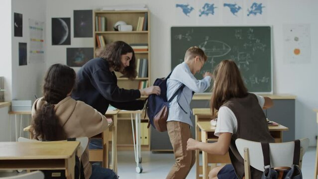 Medium long shot of teenage bullies sitting at desks in classroom greeting their classmate pushing him