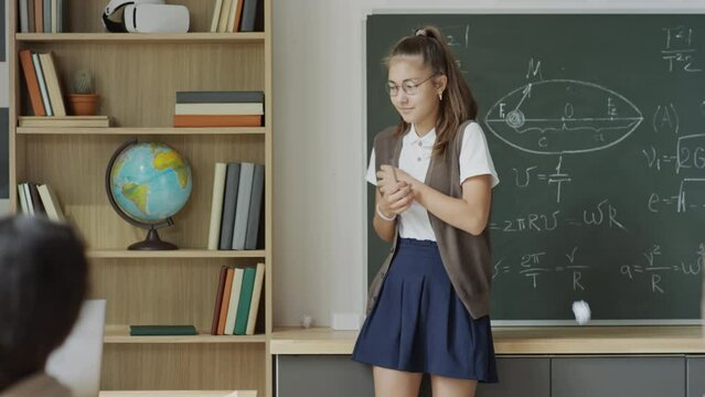 Slow motion medium long shot of teen nerd girl wearing eyeglasses standing against chalkboard in classroom suffering bullying, her classmates throwing paper balls at her