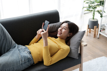 Young adorable Asian girl laying down on the sofa, using her smartphone, relaxing in her free time.