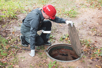 a man in overalls opened a sewer hatch and looks into a septic tank. Cleaning of sewers and drains - obrazy, fototapety, plakaty
