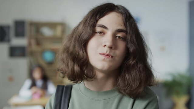 Selective focus close-up slowmo portrait of impudent teen boy with long wavy hair holding toothpick in mouth standing in classroom looking at camera