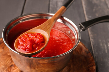 Saucepan and spoon with tasty tomato sauce on dark wooden background, closeup