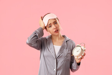 Sleepy young woman in pajamas with alarm clock on pink background