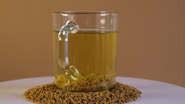 Cup of Egyptian fenugreek yellow tea, Helba or Methi Dana drink and fenugreek seeds on turntable, side view. 4K Video, Rotating. Brewed seeds used in alternative medicine.